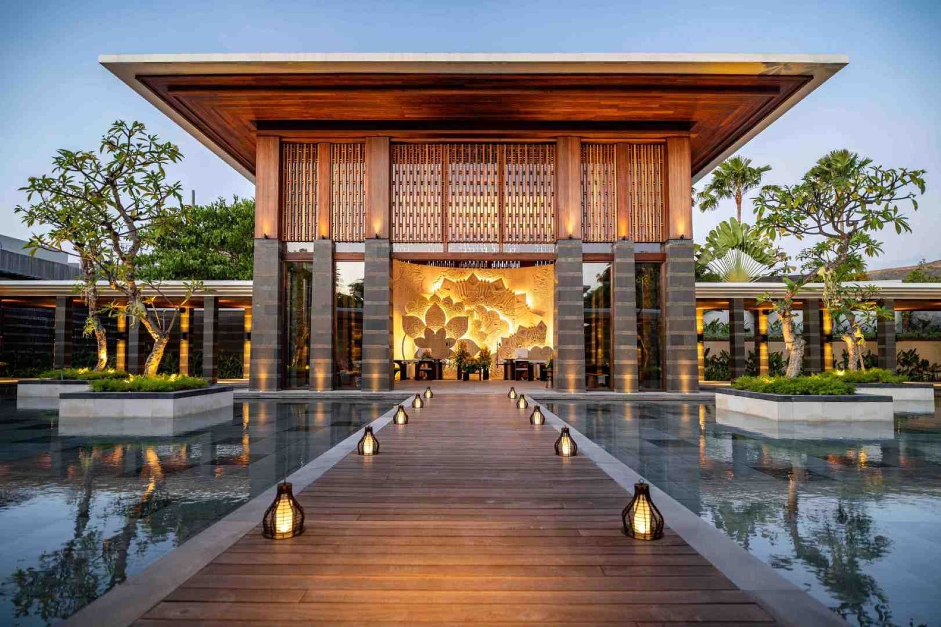 A grand wooden pavilion glows at dusk. Lanterns line a floating walkway leading to a golden floral carving. Reflecting pools frame the entrance on both sides.