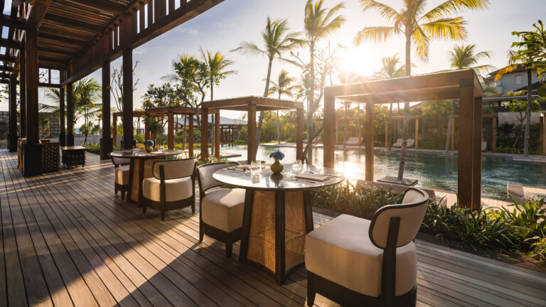 An outdoor dining terrace overlooks a lagoon pool. Rattan chairs surround marble-topped tables at sunset. Palm trees frame the golden light beyond.