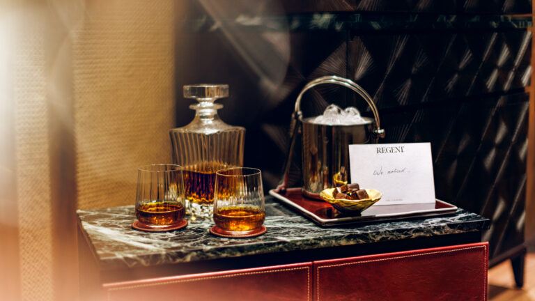 A "We noticed..." note sits beside crystal glasses. A decanter, ice bucket, and chocolates rest on dark marble. Warm lighting catches the amber drinks.