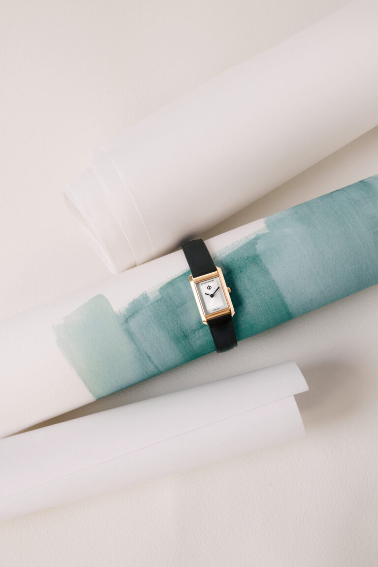 A Hampton XS in gold, laid across rolled watercolour paper. A soft teal wash bleeds across the paper beneath it. The black strap contrasts against warm, muted tones.
