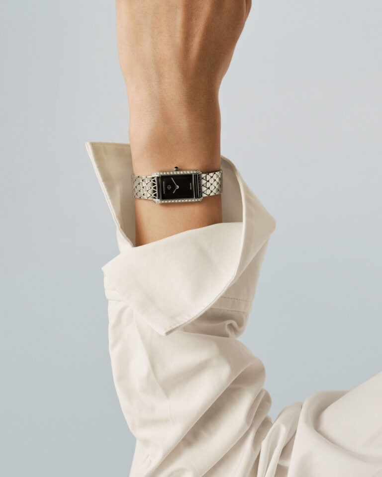 A woman’s wrist emerges from a loose white shirt sleeve. She wears a diamond-set Hampton XS with a black dial. The steel bracelet catches the light against pale skin.