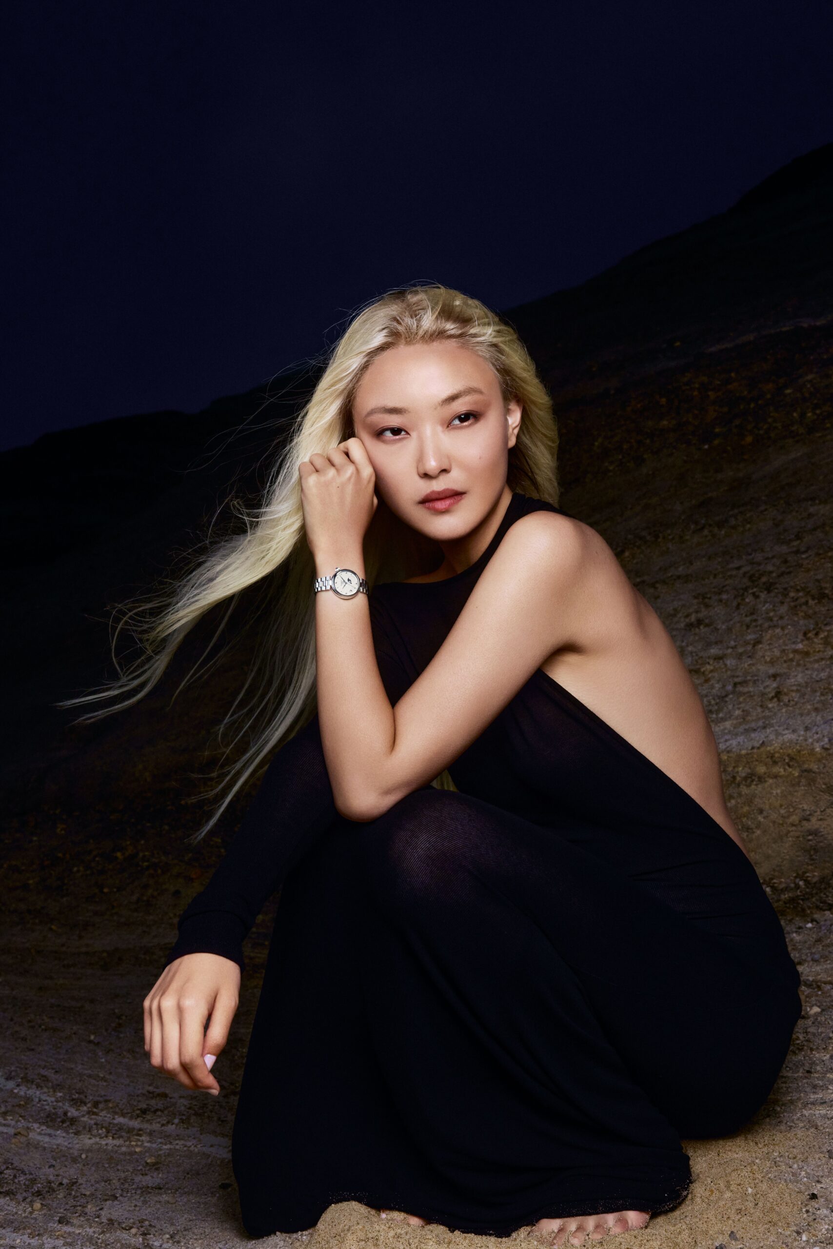 A woman crouches on rocky terrain at dusk. She wears a silver-toned Désir Moonphase on her wrist. Her hand rests near her face, watch catching the fading light.