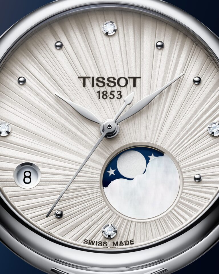 A macro shot of the silver Désir dial. Diamond hour markers ring a sunburst finish. The moonphase window glows navy with a pearl crescent moon.