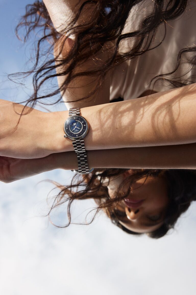 A close, mirrored shot of a wrist against pale sky. The starlit blue Désir dial sits on a steel bracelet. Windswept hair frames the reflection below.
