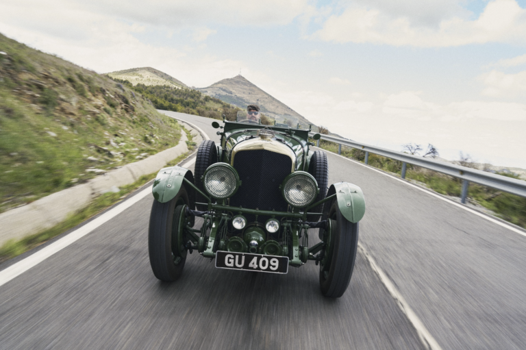 A vintage green Bentley races down a mountain road. The driver wears sunglasses behind the open cockpit. Hills roll out behind the car.